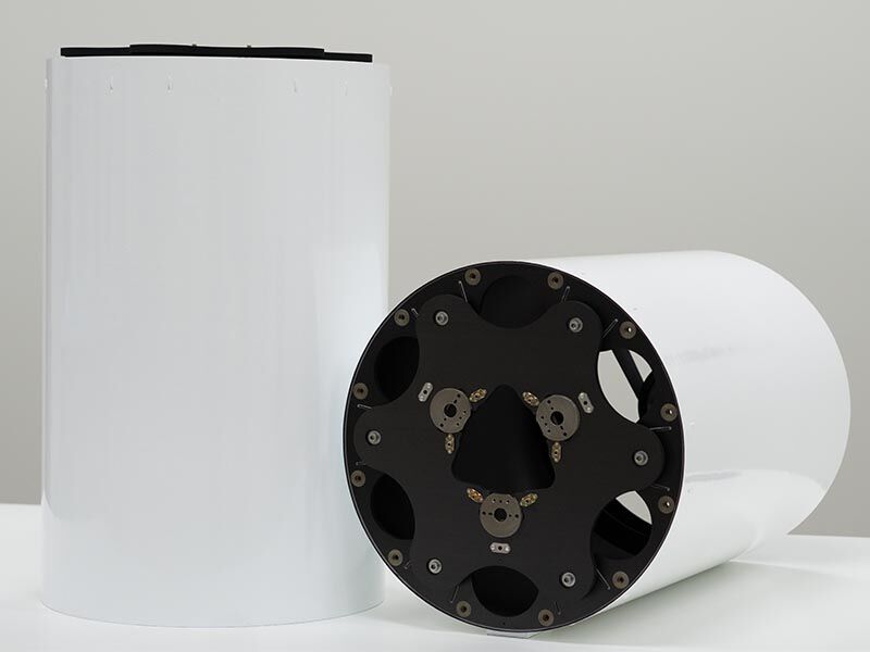 Two satellite imaging optical metering structures are shown on a white tabletop with a white background. Two large white tubes with circular side panels, made of black carbon fiber side panel with metallic inserts. One structure is laying on its side and one is place standing up.  